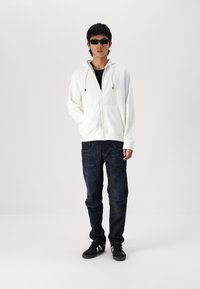 Haut à zip blanc avec cordons, t-shirt noir en dessous, jeans en denim foncé et baskets noires. Des lunettes de soleil noires complètent le look.