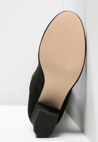 Schwarzer Wildleder-High-Heel-Schuh mit einer strukturierten Gummisohle und einer glatten beigen Innensohle. Der Absatz hat ein quadratisches Design für Stabilität.