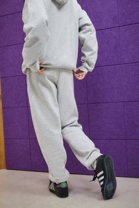 Person, der graue Sweatpants und einen Kapuzenpullover trägt, auf einem Bein steht, schwarze Adidas-Sneaker mit grünem Logo, lila geflieste Wand im Hintergrund.