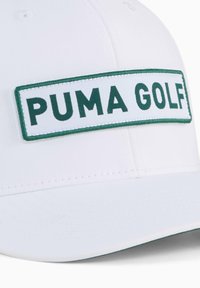 Baltas kepurė su žaliu siuvinėtų „PUMA GOLF“ emblemų. Turi struktūrizuotą dizainą ir lenktą kraštą su lygiu paviršiumi.