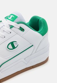 Fehér sneaker zöld díszítésekkel és perforált bőrrel. Párnázott cipőtalpa és mintázott, gumis talpa van. Champion logó látható.
