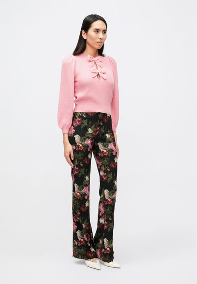 Rozā adīts džemperis ar pūkainām piedurknēm un loku pie kakla izgriezuma, kombinēts ar melnas krāsas biksēm ar ziedu rakstu un baltām smailām kurpēm ar papēdi.