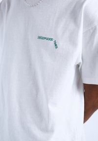 Bílá bavlněná tričko s krátkým rukávem a zeleným logem, které na levé části hrudi zobrazuje zaoblený nápis "DID IT GOOD TIMES".