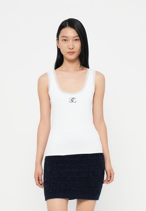 Fehér tank top áttetsző pántokkal és fekete hímzett logóval a közepén, amelyet egy texturált sötétkék mini szoknyával párosítottak.