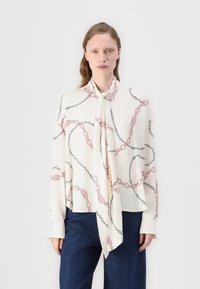 Camicia bianca con un motivo a catena in nero e rosa, dotata di un fiocco al collo e maniche lunghe. Il tessuto appare liscio e leggero.