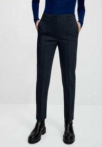 Pantaloni neri su misura con una vestibilità slim, caratterizzati da pieghe frontali, tasche laterali e una texture liscia. Abbinati a stivali neri alti fino alla caviglia.