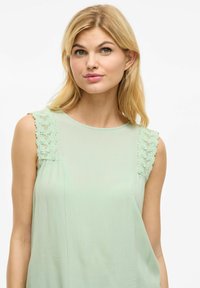 Blusa sin mangas de color verde claro con detalles de encaje en los hombros. Tejido suave y fluido con una textura lisa, que presenta un escote redondo.
