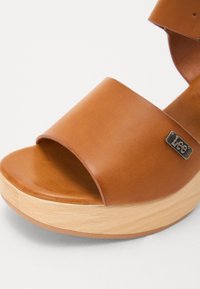 Sandal com plataforma em couro castanho, com tiras largas, bico arredondado e base de madeira. Apresenta um pequeno logo metálico na lateral.