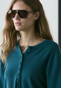 Cardigan lavorato a maglia color verde acqua con chiusura a bottoni, caratterizzato da scollatura tonda e una texture sottile. Occhiali da sole oversize scuri completano il look.