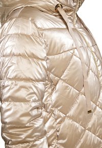 Giacca a puffo beige chiaro con finitura lucida, motivo a rombi, chiusura con zip e cappuccio regolabile con cordino. Accentuata da hardware dorato.