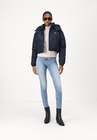 Marineblå, kort pufferjakke med hette, lys beige genser og lyseblå skinny jeans. Svarte høyhælte ankelstøvler, solbriller og ingen synlige mønstre.