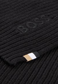Svart ribbestrikket stoff med preget "BOSS"-logo og et lite merke med svarte, hvite og beige striper.