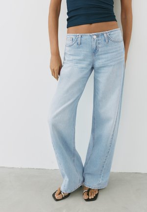 Personne portant un jean large bleu clair, des sandales noires à bout carré et un débardeur court foncé, debout sur un sol gris.