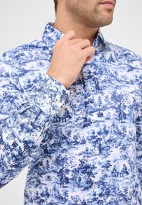 Man die de kraag van een witte overhemd met gedetailleerde blauwe toileprint aanpast, met bomen, mensen, boten en gebouwen.