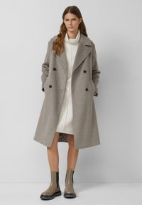 Dwurzędowy płaszcz z mieszanki wełny w kolorze szarym z dużymi guzikami, klapami z wycięciem i kieszeniami; noszony na jasnej sukience i botkach do kostek.