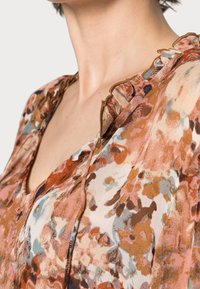 Blusa de tejido ligero y transparente con un estampado floral, que presenta suaves volantes alrededor del escote. Los colores incluyen tonos de rosa, marrón y azul.