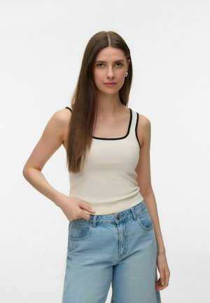 Jeune femme aux longs cheveux bruns portant un débardeur crème avec finition noire et un jean bleu clair, debout devant un fond blanc.