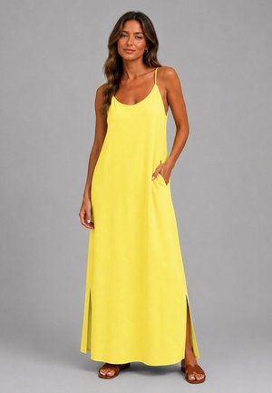 Femme portant une robe maxi jaune sans manches avec des fentes latérales et des sandales marron, debout devant un fond gris uni.
