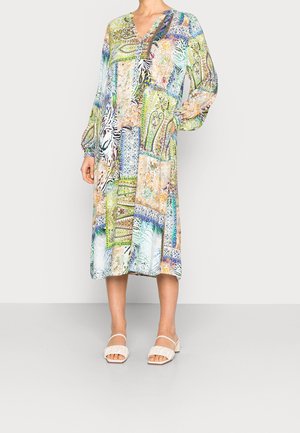 Vrouw draagt een kleurrijke midi-jurk met patronen, lange mouwen en een v-hals, gecombineerd met witte sandalen met hak.