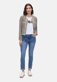Veste texturée beige et noire au design court, portée sur un t-shirt graphique blanc et un jean bleu, associée à des baskets blanches.