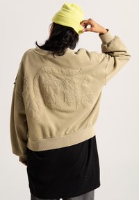 Felpa khaki con vestibilità oversized, con "Vans" ricamato in una texture a contrasto sul retro. Maglietta nera sotto.