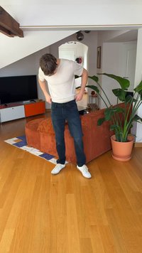 Blugi închiși la culoare și un tricou deschis la culoare purtate de o persoană care își ajustează hainele într-o cameră cu podea din lemn și o plantă verde în apropiere.