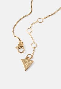 Gold chain necklace with a triangular charm displaying "GUESS" and a question mark. Features a lobster clasp and circular links.