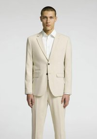 Beżowy garnitur o dopasowanym kroju, marynarka zapinana na jeden guzik, z klapami, oraz dopasowane spodnie. Biała koszula z mankietami na guziki.