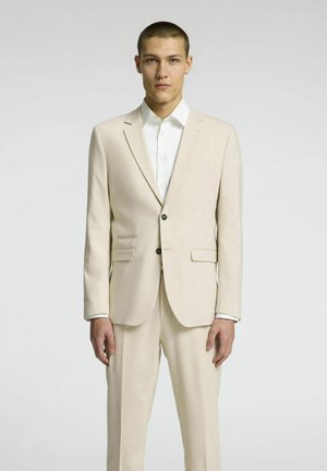 Beige jakkesæt med skræddersyet pasform, single-button jakke, notch-lapel og matchende bukser. Hvid skjorte med knapmanchetter.