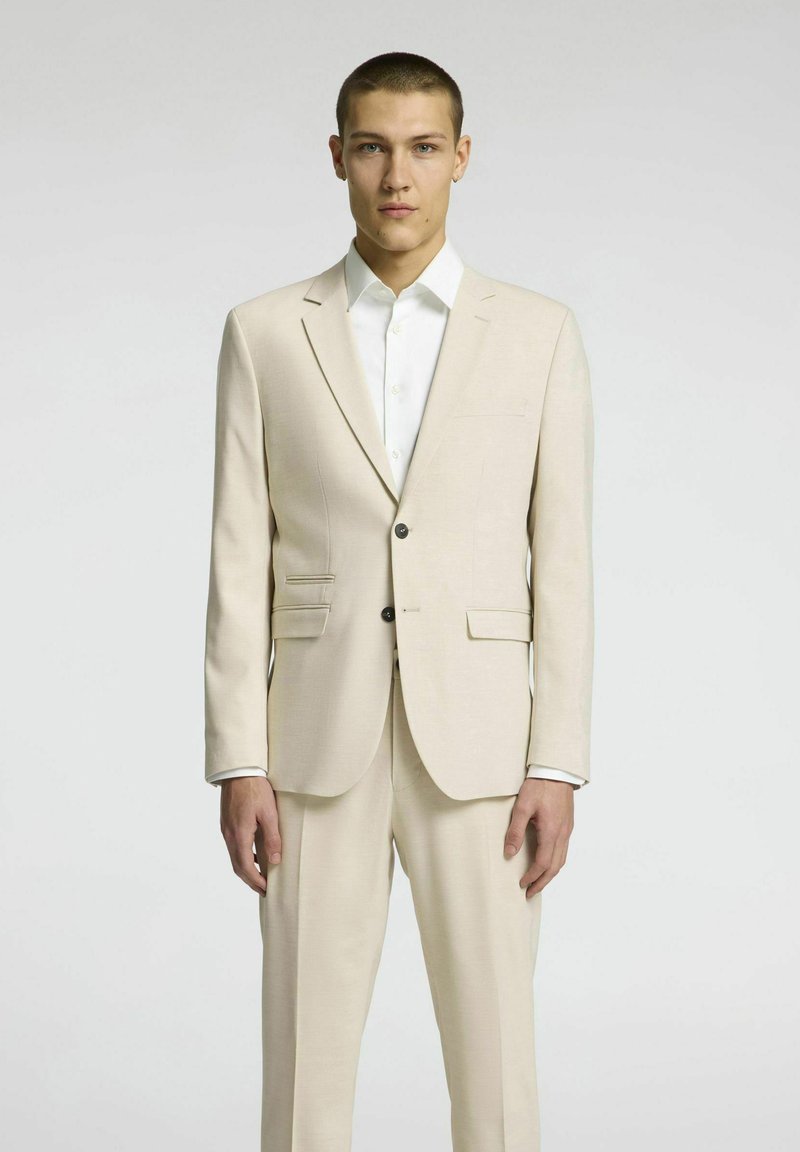 Beżowy garnitur o dopasowanym kroju, marynarka zapinana na jeden guzik, z klapami, oraz dopasowane spodnie. Biała koszula z mankietami na guziki.