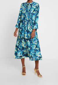 Langes, florales Kleid in Blau-, Grün- und Gelbtönen. Verfügt über einen runden Ausschnitt, lange Ärmel und plissierte Details. Getragen mit gelben Riemchenschuhen.