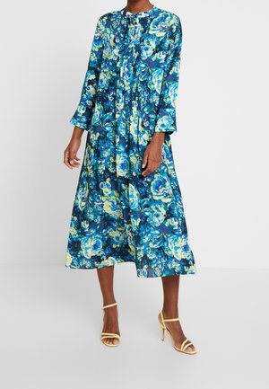 Robe longue à fleurs dans des tons de bleu, de vert et de jaune. Présente un col rond, des manches longues et des détails plissés. Portée avec des talons jaunes à lanières.