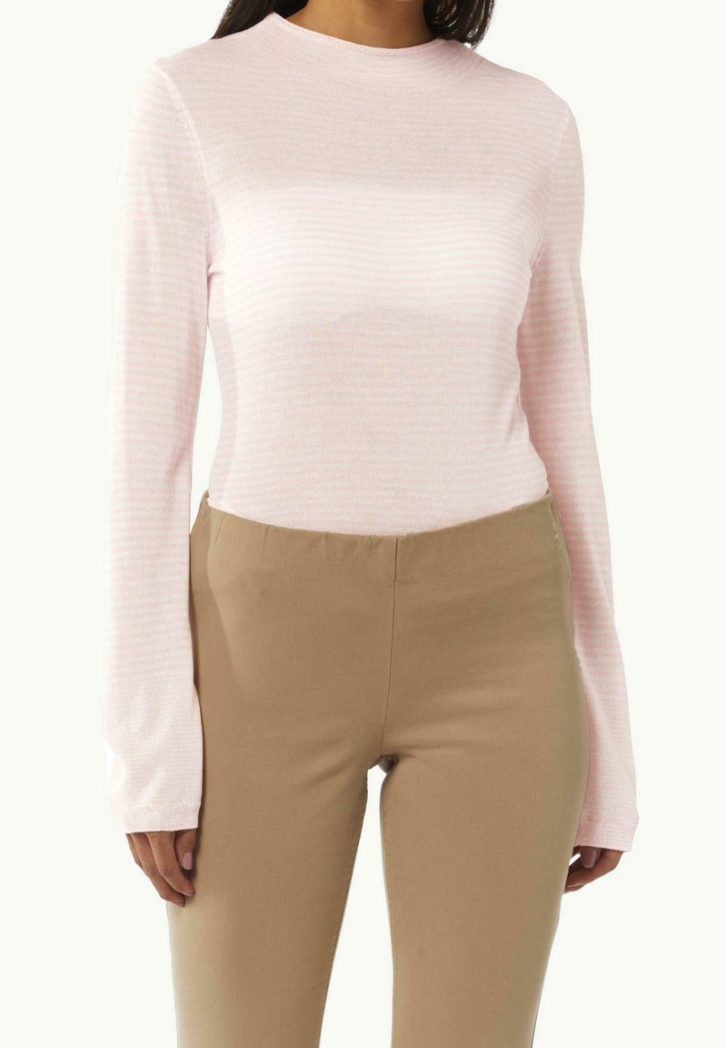 Femme portant un haut ajusté à manches longues rose clair et un pantalon beige taille haute, debout devant un fond blanc uni.