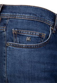 Poche d'un jean en denim bleu foncé avec des coutures orange visibles et un petit logo carré en métal portant les lettres "KL" sur la poche à monnaie.