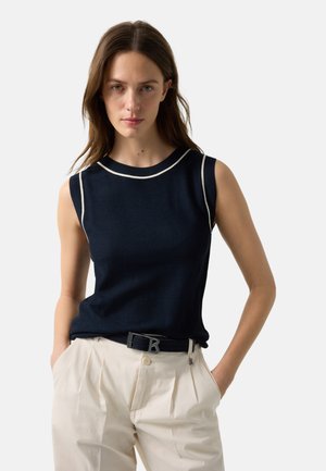 Femme portant un haut sans manches bleu marine avec des finitions blanches et un pantalon beige taille haute, les mains dans les poches, debout devant un fond uni.