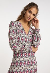 Rochie cu mânecă lungă dintr-un material ușor, având un model geometric în formă de diamant în culorile roșu, roz și negru pe un fundal crem, cu decolteu în V.