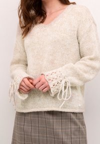 Sweter w kolorze kremowym, z dekoltem w serek, wykonany z dzianiny, z luźnymi rękawami ozdobionymi koronką i sznurkami przy mankietach. Nosi się go na spódnicy w kratę.