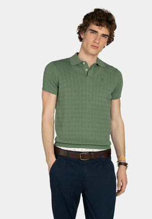 Joven con cabello rizado que lleva una camisa polo verde de punto trenzado, pantalones azul marino, cinturón marrón y muñequeras, de pie con una mano en el bolsillo.