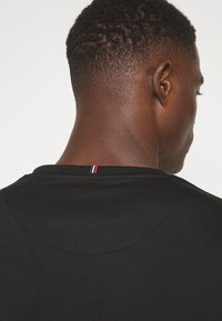 Schwarzes T-Shirt aus Baumwolle mit runder Halslinie. Verfügt über einen kleinen roten, weißen und blauen Akzent am hinteren Halsausschnitt. Glatte Textur.