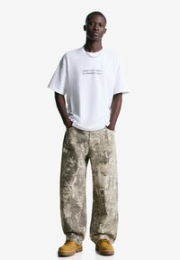 Witte katoenen T-shirt met bedrukte tekst, gecombineerd met gestreepte beige cargobroek en bruine veterschoenen. Effen achtergrond.
