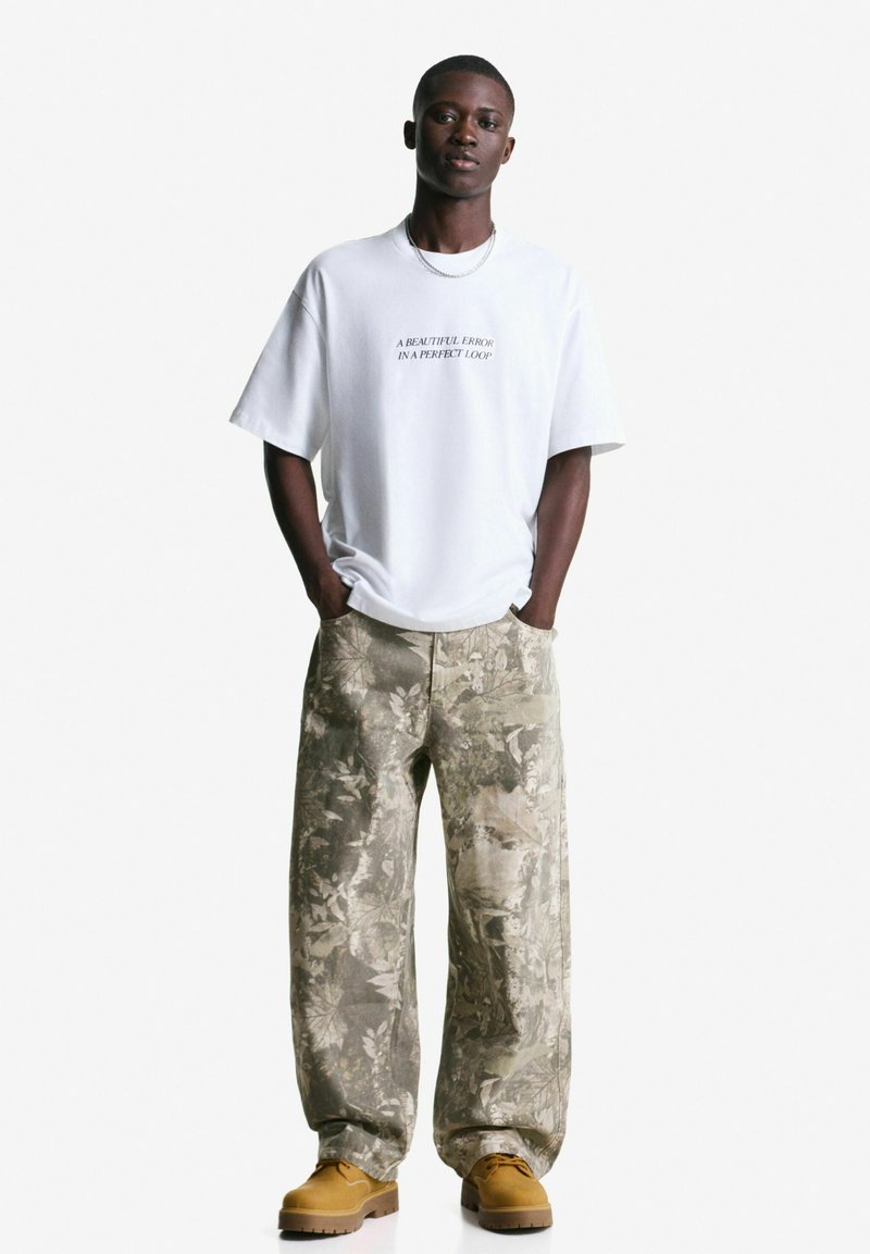 Witte katoenen T-shirt met bedrukte tekst, gecombineerd met gestreepte beige cargobroek en bruine veterschoenen. Effen achtergrond.