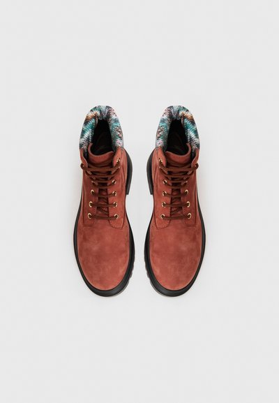 Brown suede ankle boots with black rubber soles, metal eyelets, and brown laces. Patterned fabric trim at the top adds texture.