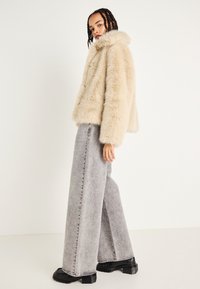 Faux furjakke i beige med høy krage, parret med grå denimbukser med vide ben og kraftige svarte sko, satt mot en nøytral bakgrunn.