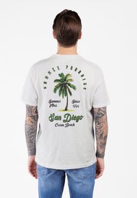 Hombre con tatuajes en ambos brazos que lleva una camiseta blanca con una palmera verde y el texto "Verano Paraíso San Diego Ocean Beach" en la parte posterior.