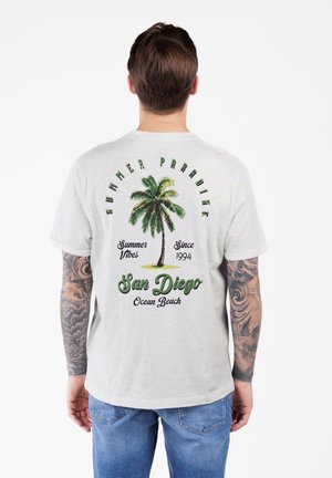 Mann mit Tattoos an beiden Armen trägt ein weißes T-Shirt mit grünem Palmenmotiv und dem Text "Sommerparadies San Diego Ocean Beach" auf dem Rücken.