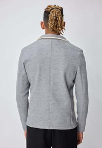 Homme aux dreadlocks blonds attachées à l'arrière, portant une veste cintrée gris clair à manches longues et un pantalon noir, vu de dos sur un fond uni.