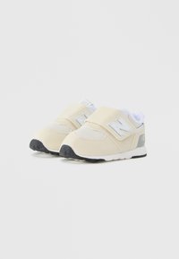 Sneakers beige con tomaia in mesh e suede, strap con velcro e suola in gomma nera. Presentano un logo New Balance bianco sul lato.