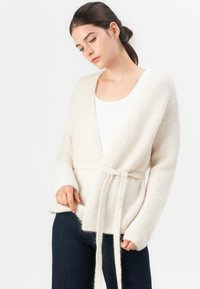 Ung kvinne som har på seg en hvit, fluffy belted kardigan over en hvit topp og mørke jeans, står mot en enkel hvit bakgrunn.