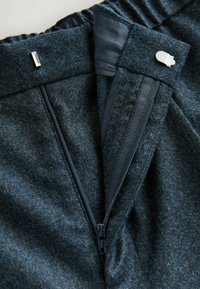 Dark grey wool trousers featuring a zippered fly, smooth satin interior, elastic waistband, and metal clasp. Close-up view of fastening details.