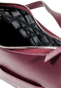 Borsa a tracolla testurizzata color bordeaux con chiusura a zip. L'interno foderato in nero presenta etichette del marchio ripetute. Le maniglie sono lisce e robuste.